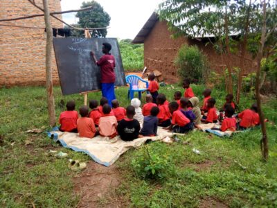 Ugandan woman teaching a group of young children outside in the grass with a blackboard and blue chalk